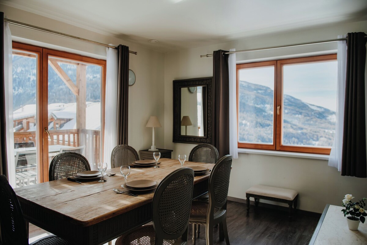 Un comedor con una mesa de madera, sillas y vistas a montañas a través de amplios ventanales.