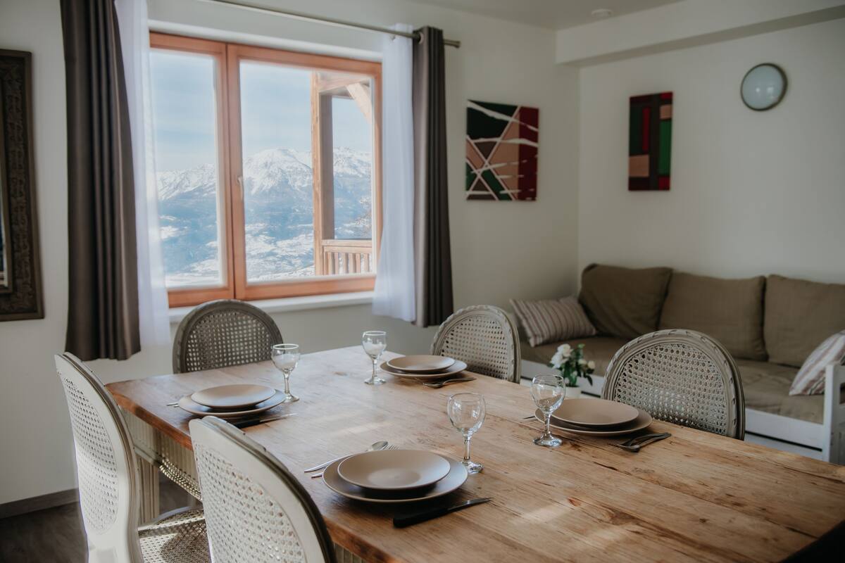A dining area with a wooden table set for four, chairs, a view of mountains through a window, and a sofa in the background.