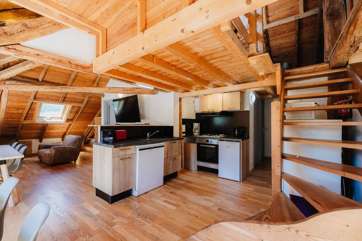 A cozy attic-style room with a wooden ceiling, a small kitchenette, a dining area with a table and chairs, and a staircase.