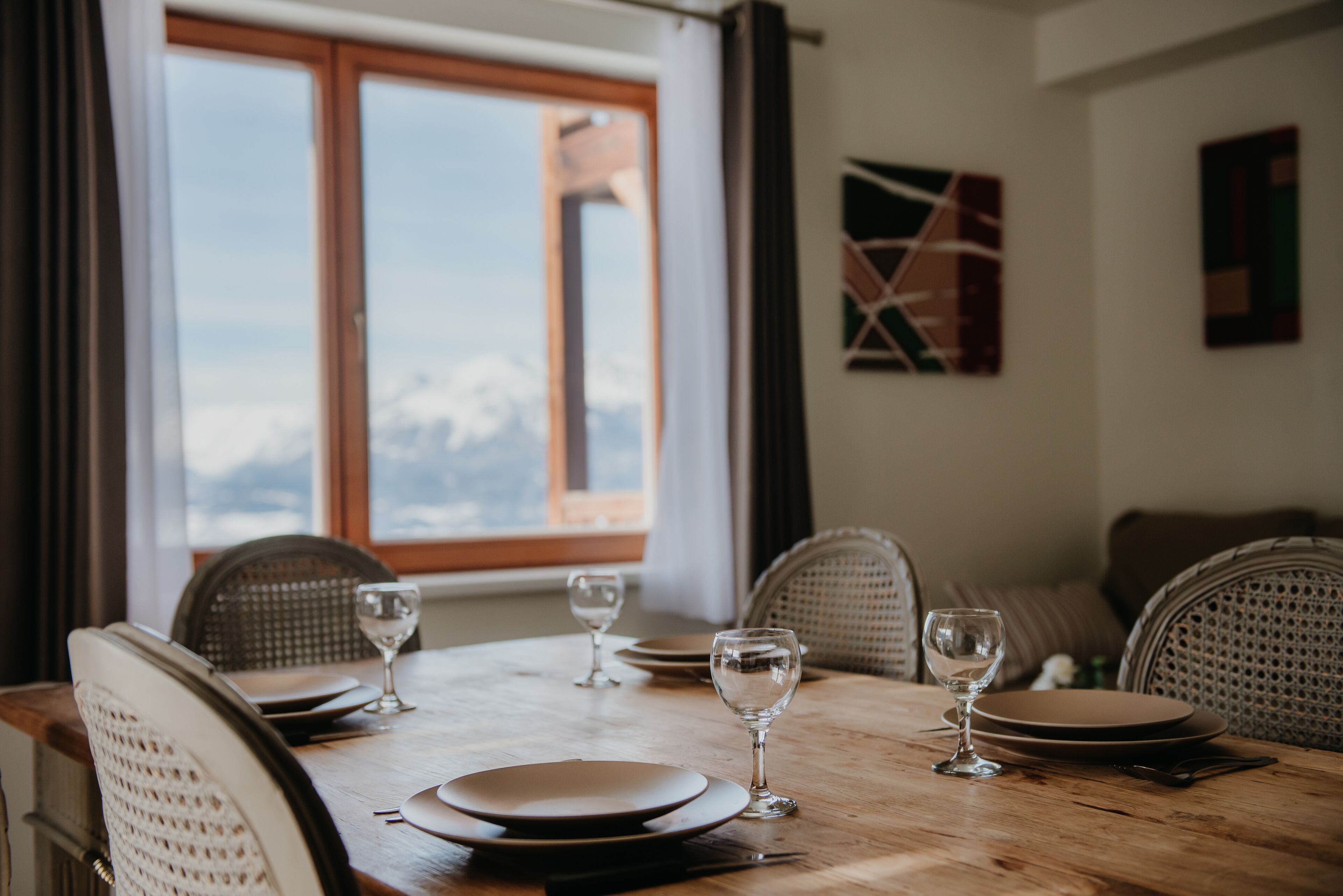 A dining table set with plates and wine glasses, a window with a view of mountains, and wicker chairs.