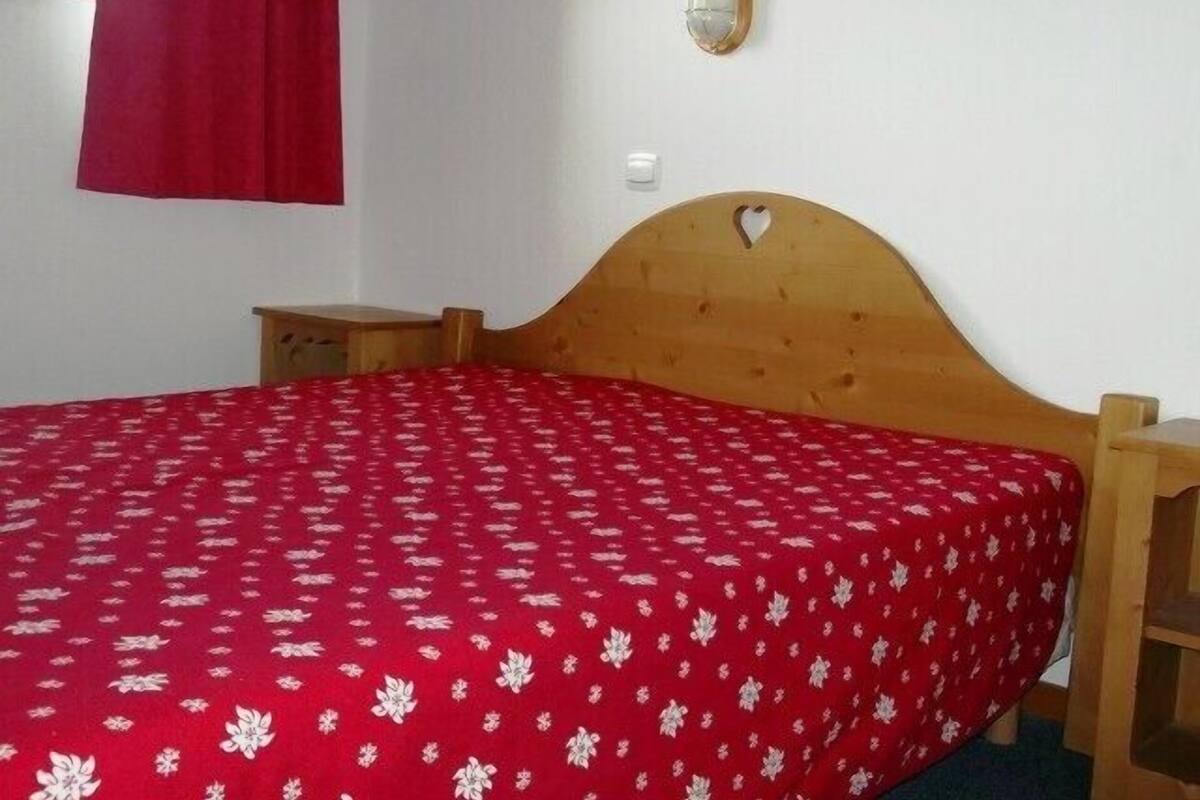 Ein Schlafzimmer mit einem hölzernen Bett, einer roten Blumenbettdecke, einem Nachttisch, einer Wandleuchte und einem Fenster mit rotem Vorhang.