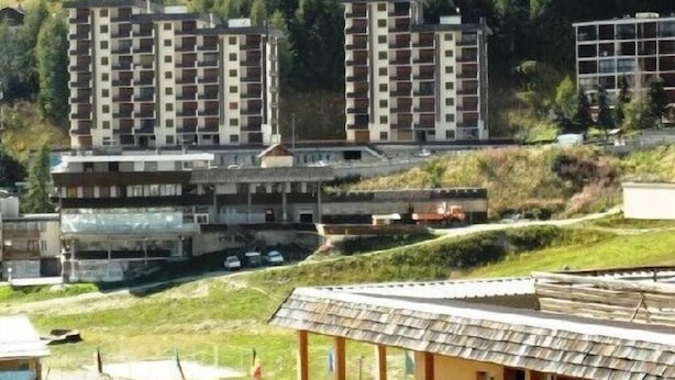A multi-story building with balconies, a parking area, and a small structure with a tiled roof.