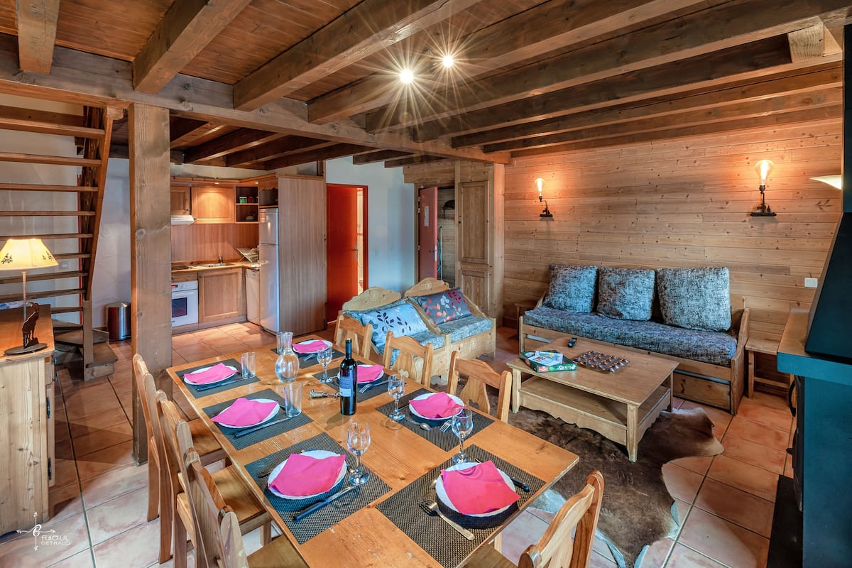 A rustic wooden interior with a dining area set for four, a sofa with cushions, and a kitchen in the background.