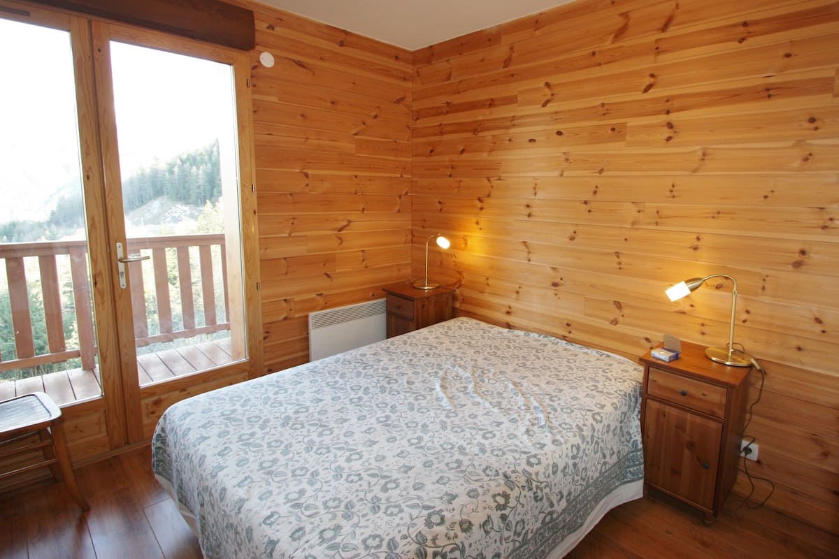 A wooden cabin bedroom with a bed, bedside lamps, a nightstand, and a sliding glass door leading to a balcony.