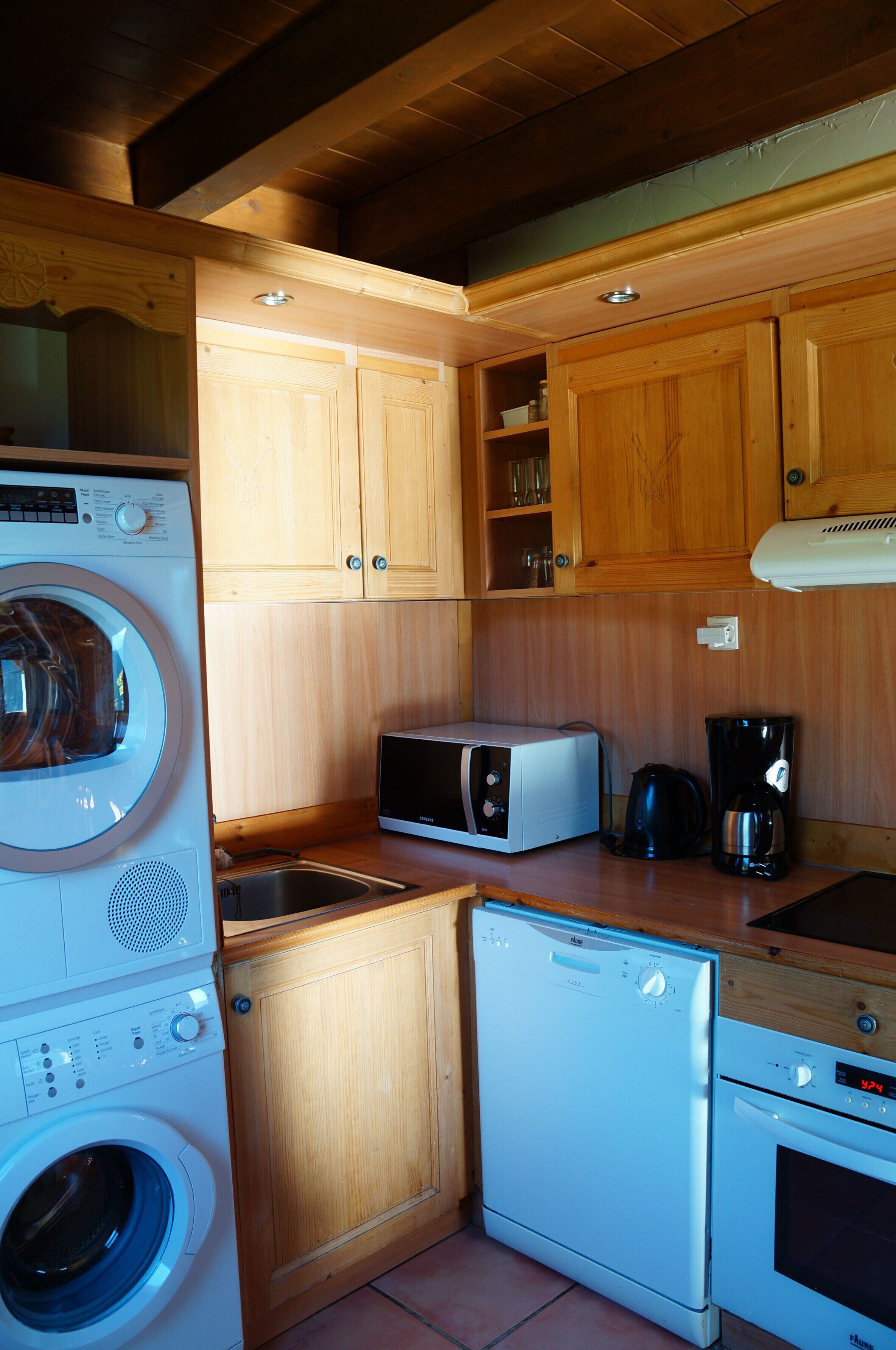 A compact kitchen with wooden cabinets, a washing machine, a microwave, a coffee maker, and a dishwasher.