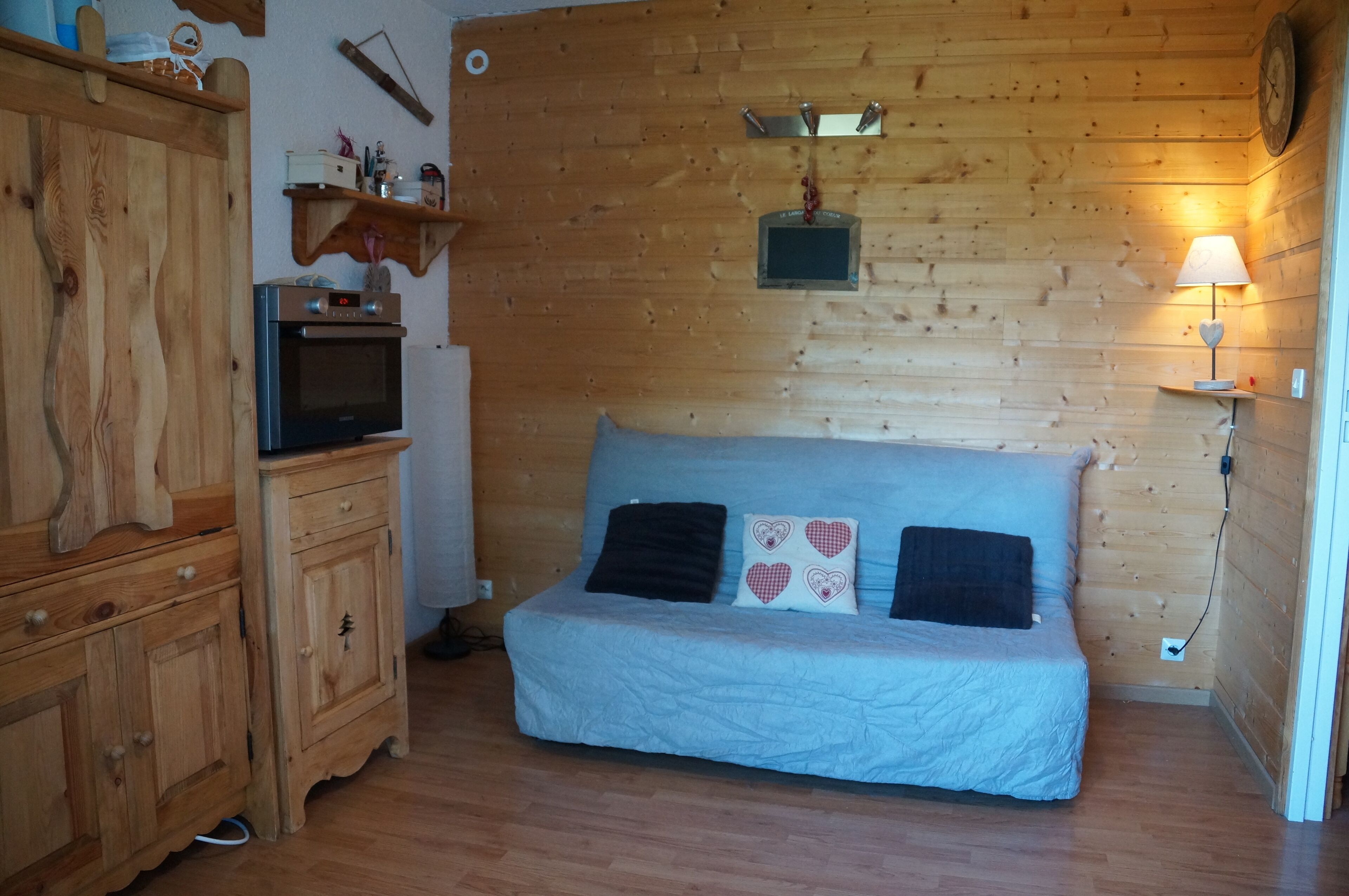A cozy room with a wooden floor, a bed with heart-patterned pillows, a wooden cabinet, and a wall-mounted TV.