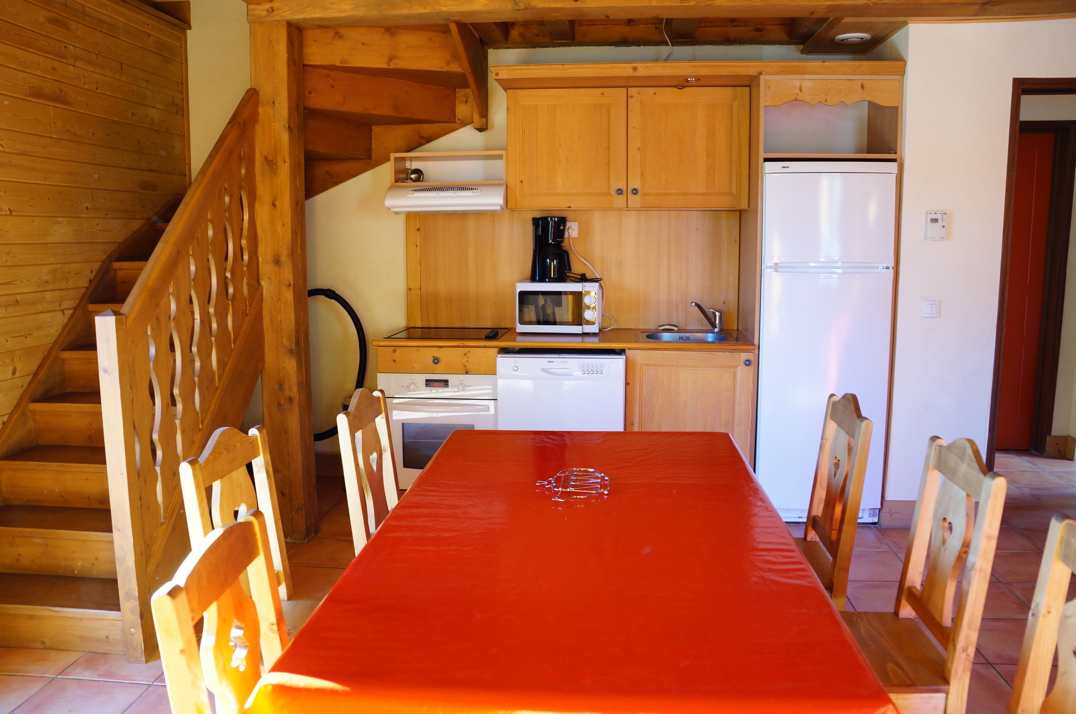 A wooden staircase leading to a dining area with a red table and chairs. A kitchen with wooden cabinets, a microwave, and a refrigerator is visible in the background.