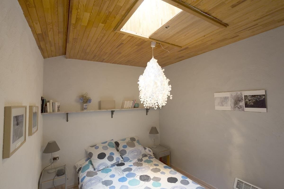 Een slaapkamer met een houten plafond, een bed met een stippenbedover, nachtkastjes met lampen, een plank met boeken en een vaas, en een dakraam.