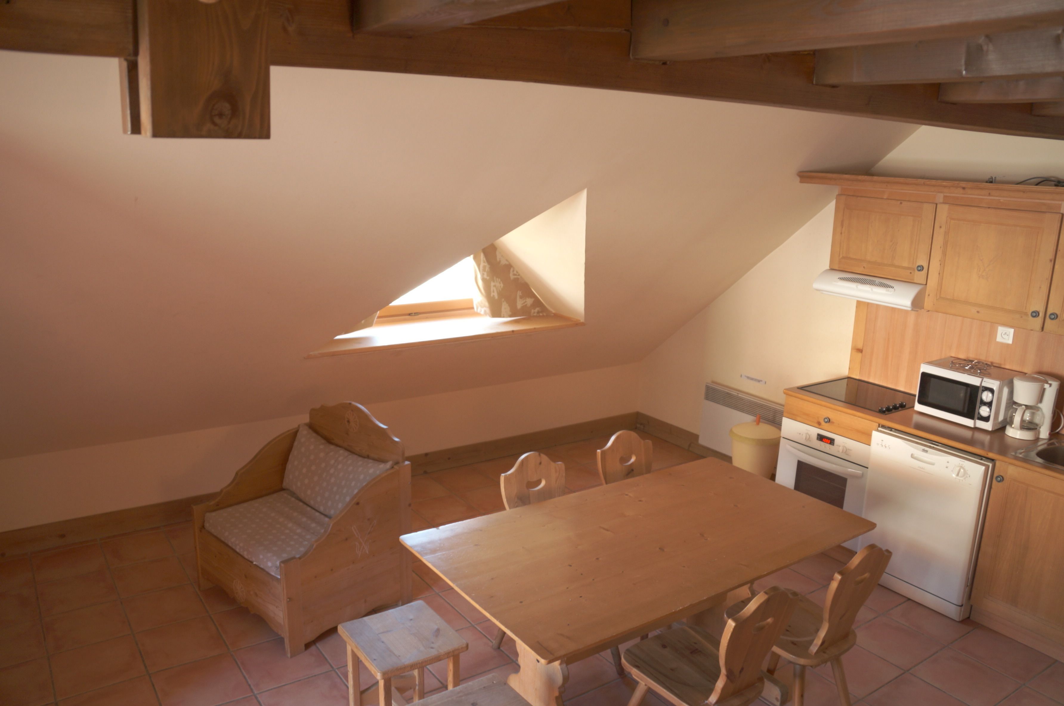 A small, cozy kitchen with a wooden table and chairs, a built-in microwave, and a small oven. There is a window providing natural light.