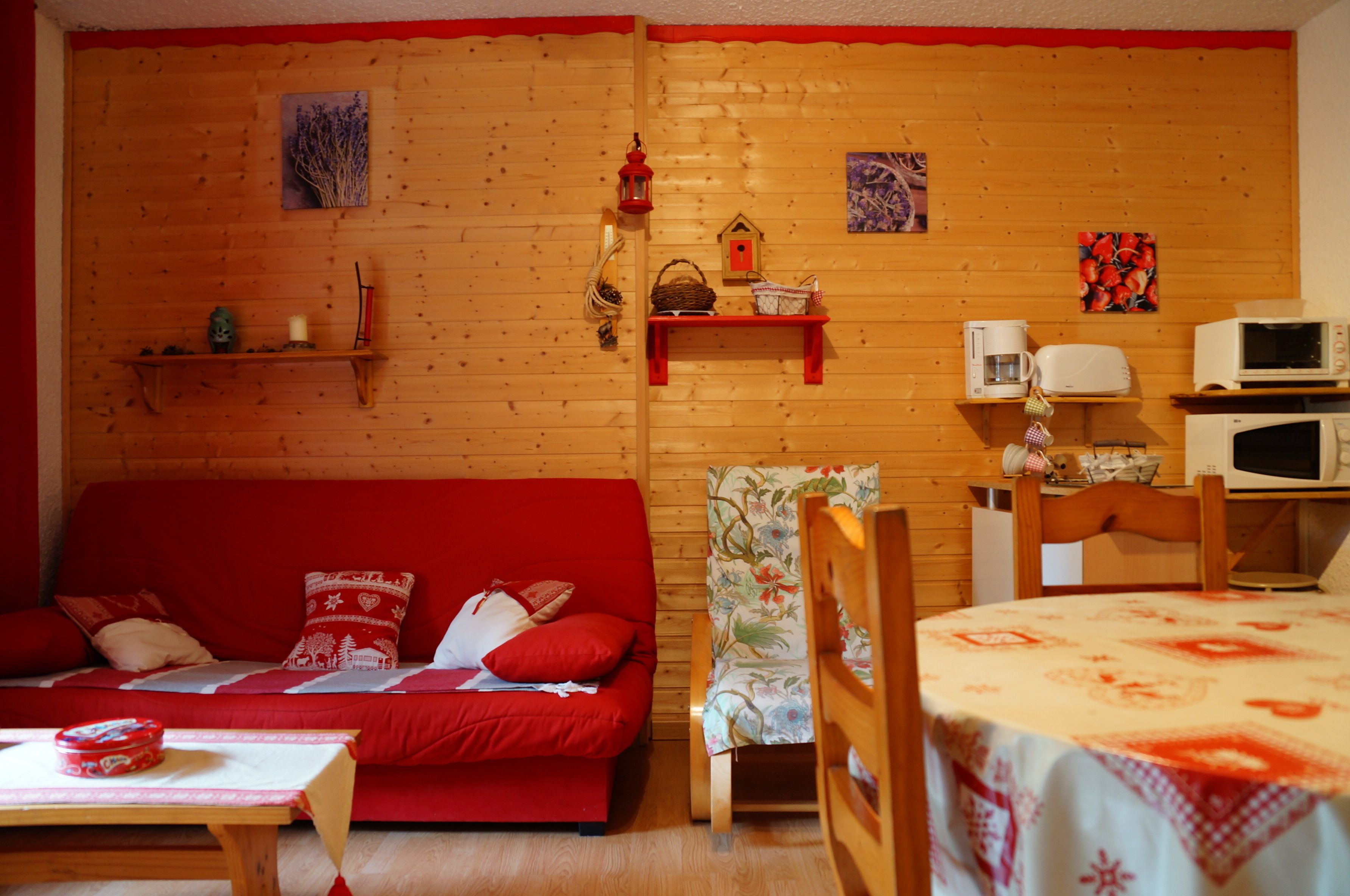 A cozy room with a wooden wall, a red sofa, a floral armchair, a dining table, and a microwave.