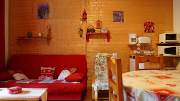 A cozy room with a wooden wall, a red sofa, a floral armchair, a dining table, and a microwave.