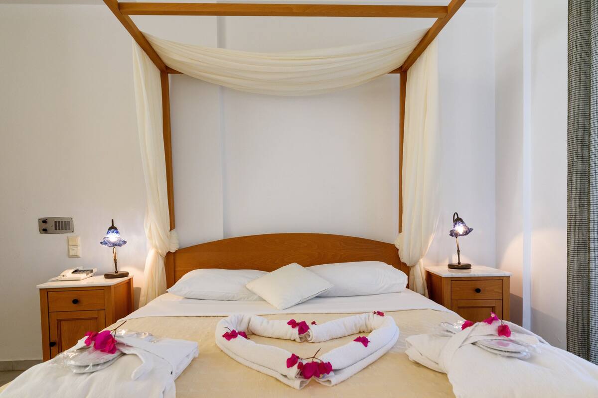 A neatly made bed with white linens, a wooden headboard, and a canopy. There are two bedside tables with lamps, a nightstand, and a wall-mounted telephone.
