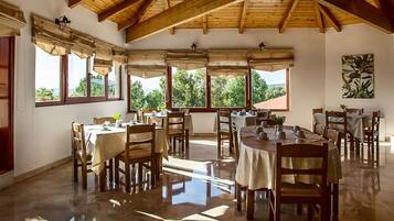 Un comedor con varias mesas y sillas, ventanas amplias con cortinas y techo de madera.