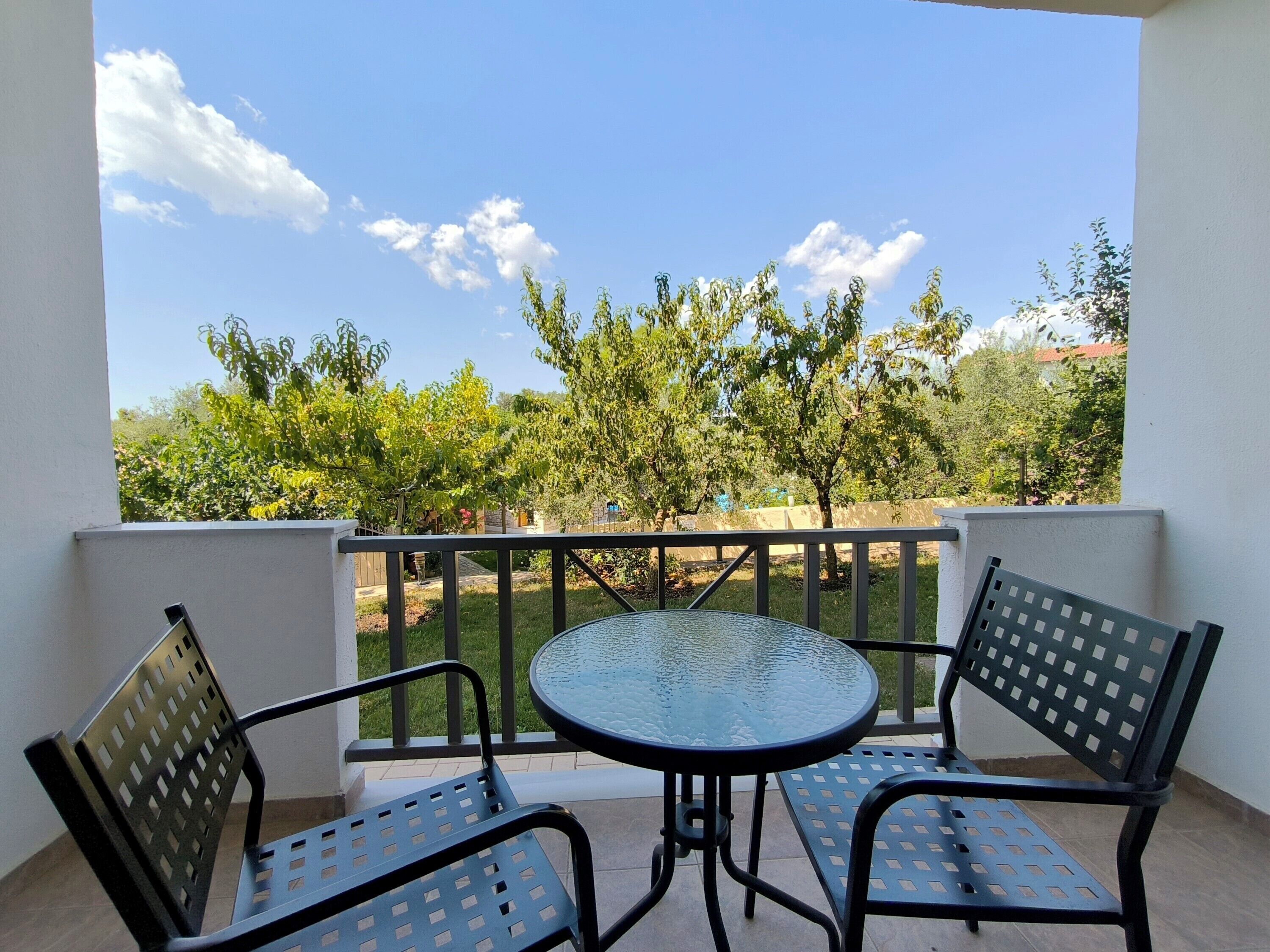 Un balcon avec une table ronde en verre et deux chaises en métal, donnant sur un jardin avec des arbres et un ciel dégagé.