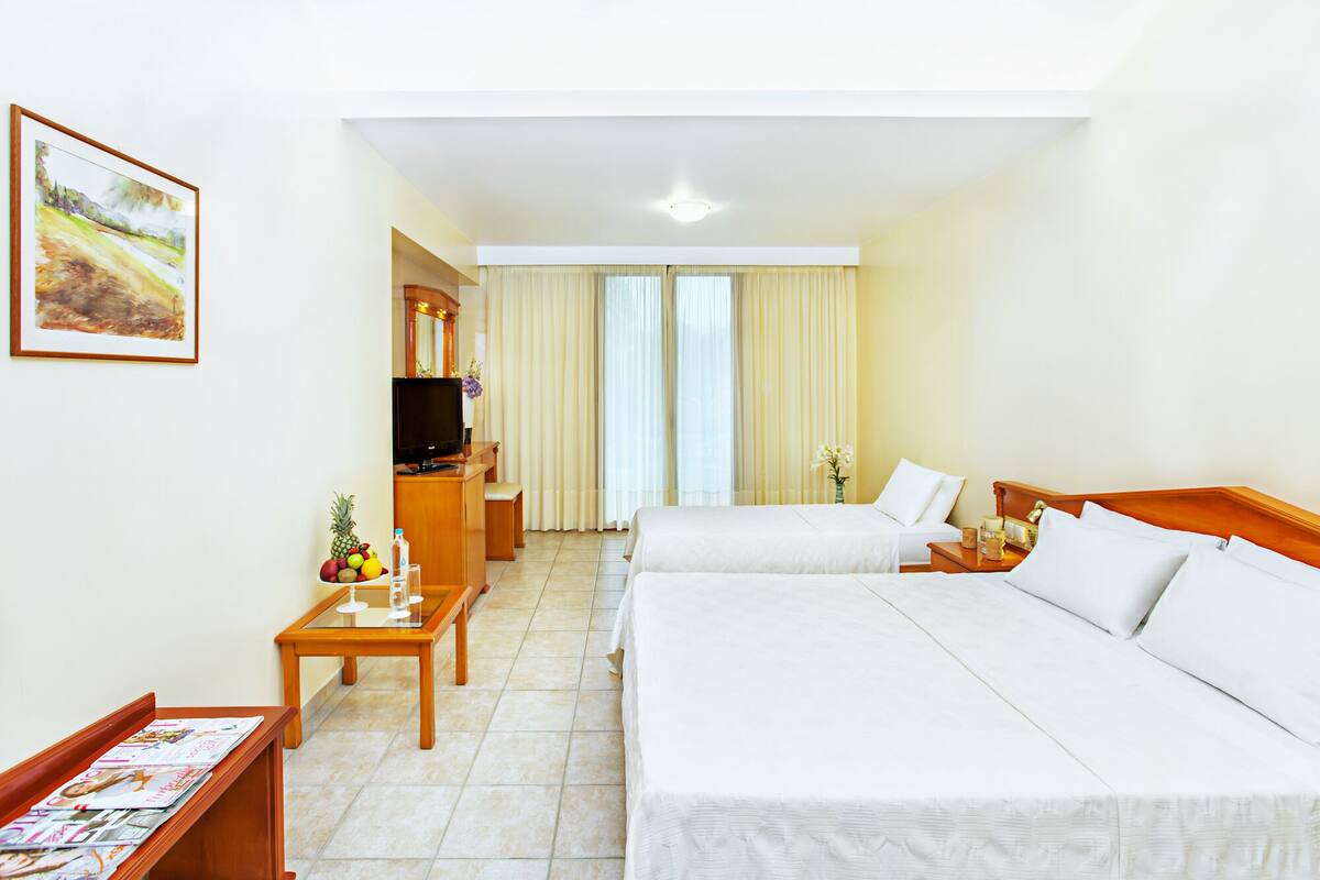Habitación de hotel con dos camas, un escritorio con computadora, un televisor y una mesa de comedor con fruta y revistas.