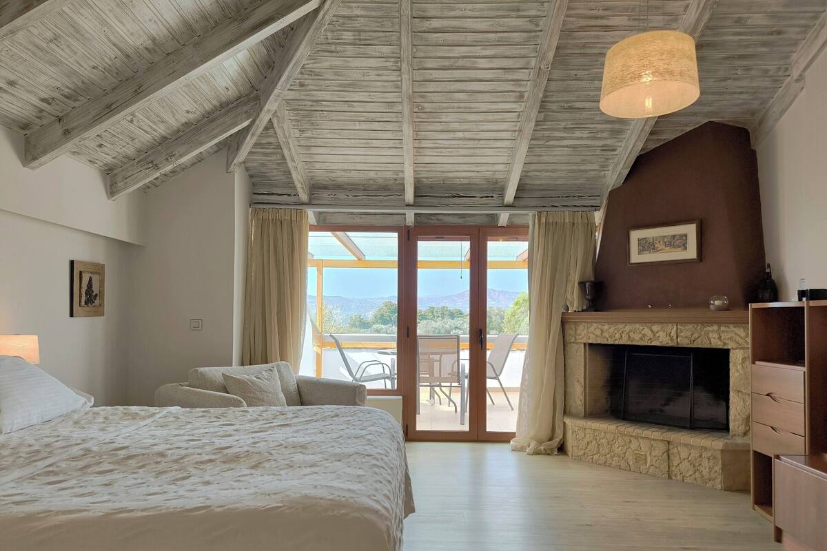Un dormitorio con una cama grande, una chimenea, vistas a través de un ventanal y un techo con vigas a la vista.