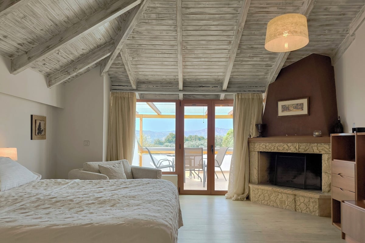 A bedroom with a large bed, a fireplace, a view through a glass door, and a ceiling with exposed beams.