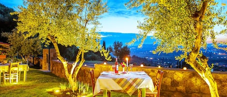 Un espace de restauration en plein air avec une table dressée pour deux personnes, entouré d’oliviers et bordé d’un mur de pierre, offrant une vue sur un paysage pittoresque.