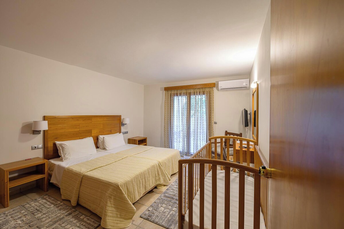 Habitación de hotel con cama de cabecera de madera, una mesita de noche con lámpara, una pequeña mesita auxiliar de madera, una ventana con cortinas, una silla de madera, un escritorio y una silla.