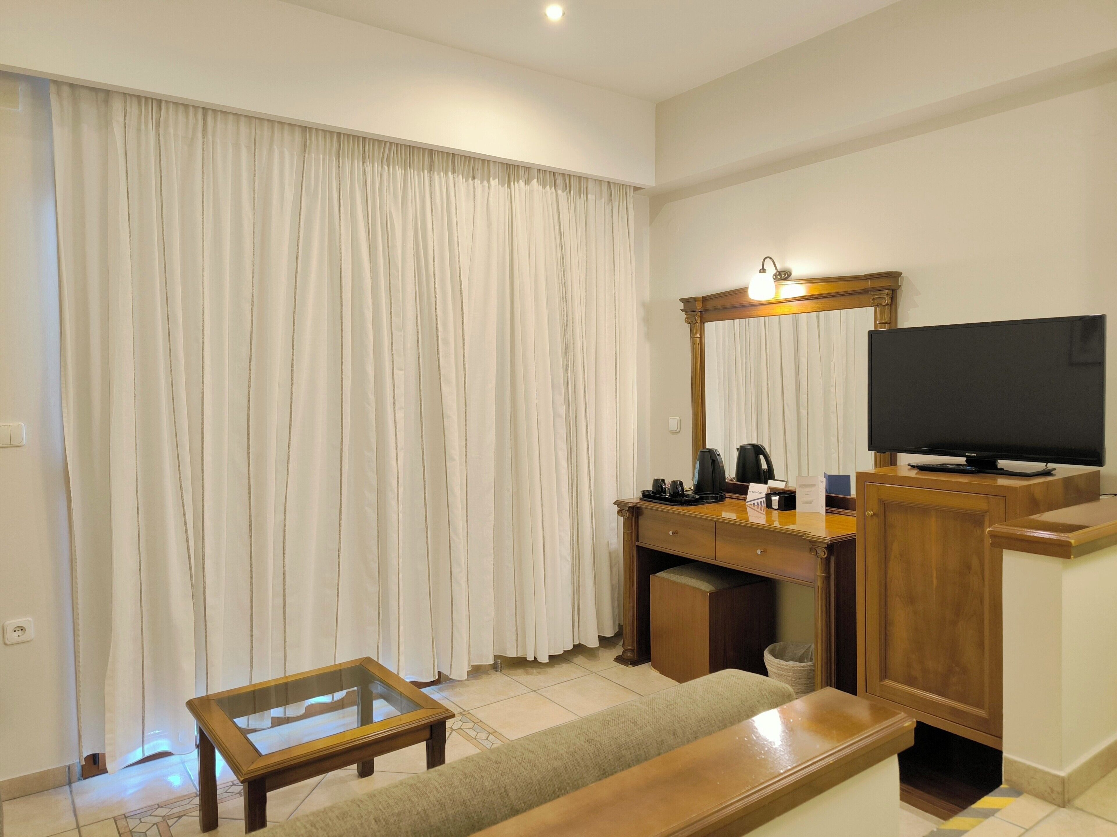 A hotel room with a wooden desk, a television, a sofa, and a large window with white curtains.