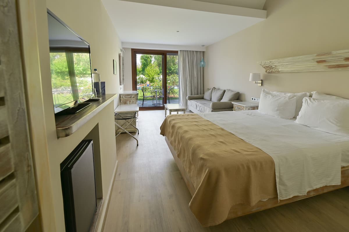 Habitación de hotel con una cama grande, televisión, escritorio y balcón con vistas a zonas verdes.