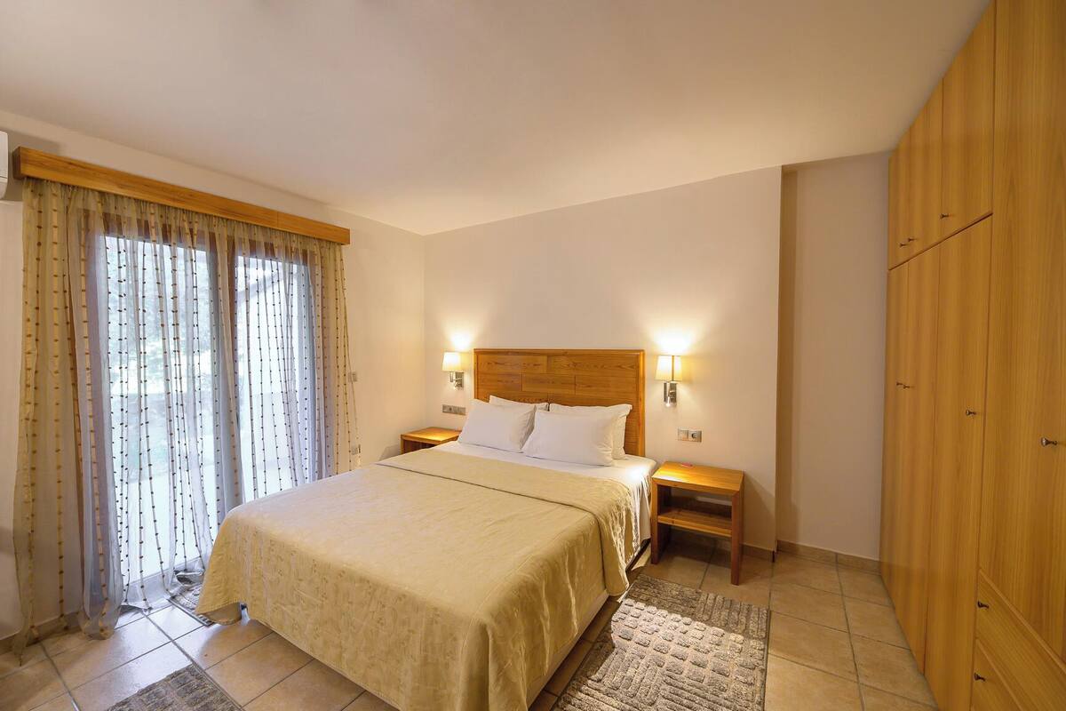 Una habitación de hotel con cama, dos lámparas de noche, un armario de madera y una ventana con cortinas.