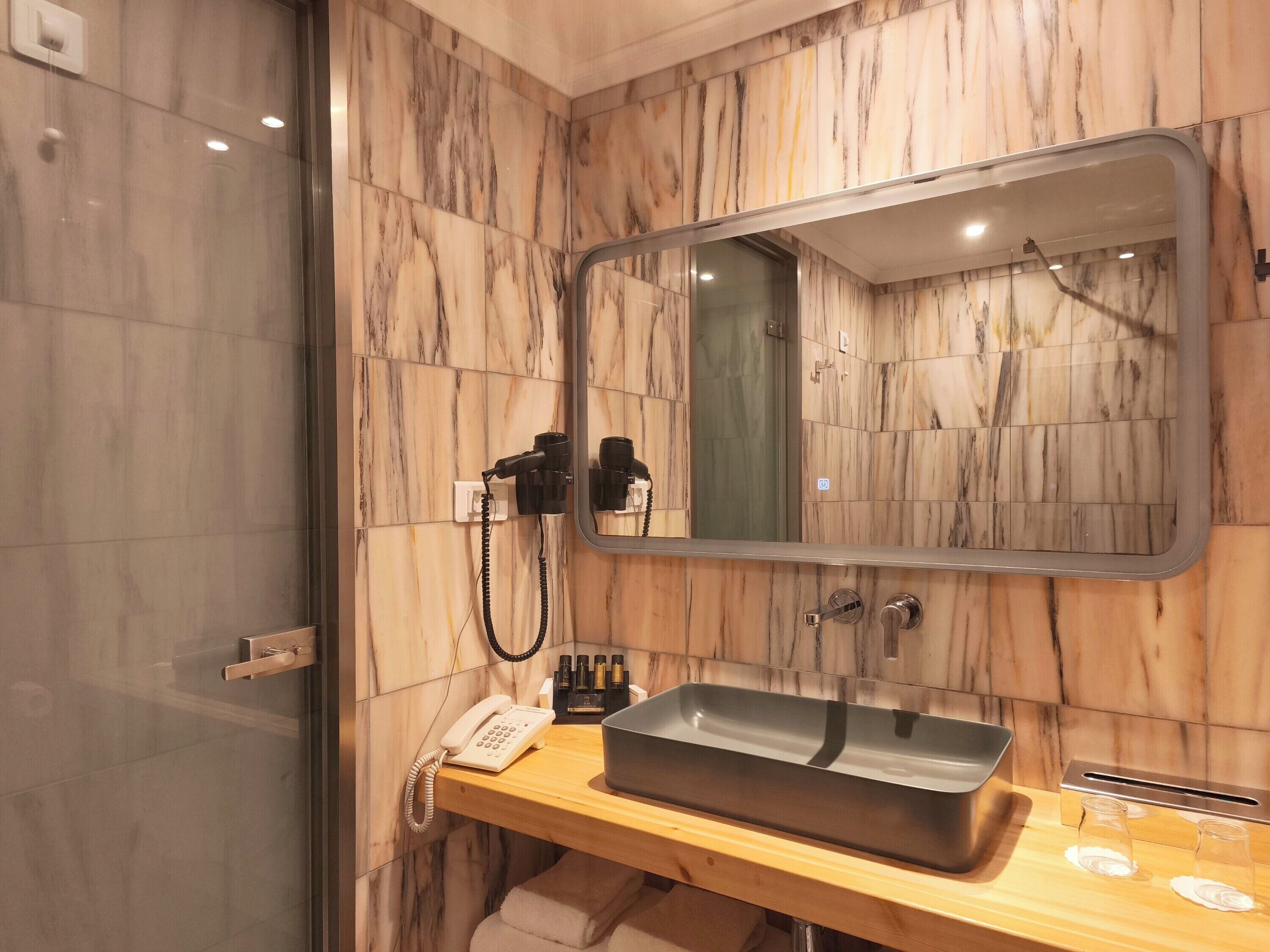 A bathroom with a marble wall, a sink with a basin, a mirror, a hairdryer, and a telephone.