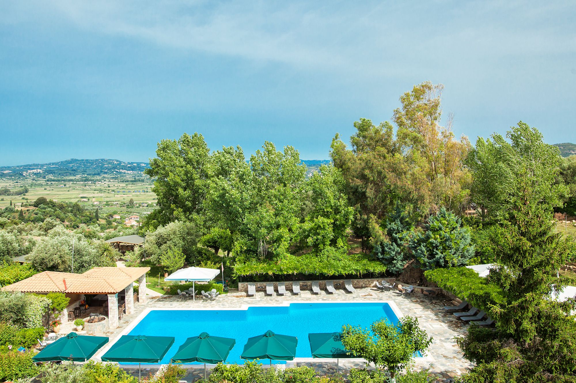 A pool area with lounge chairs and umbrellas, surrounded by trees and a view of the countryside.