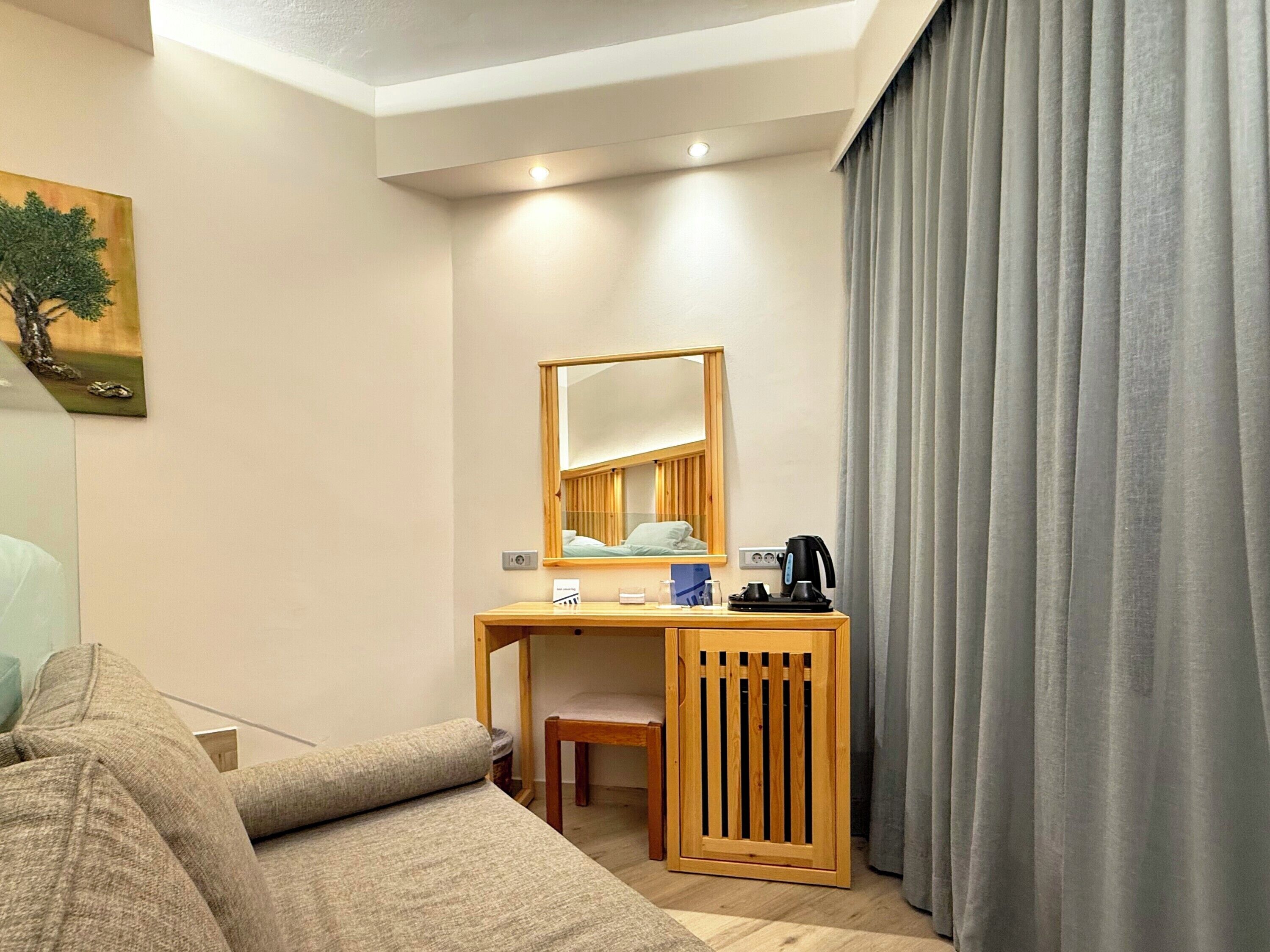 Una habitación de hotel con cama, escritorio con silla, una mesita, espejo, cafetera y ventana con cortinas.