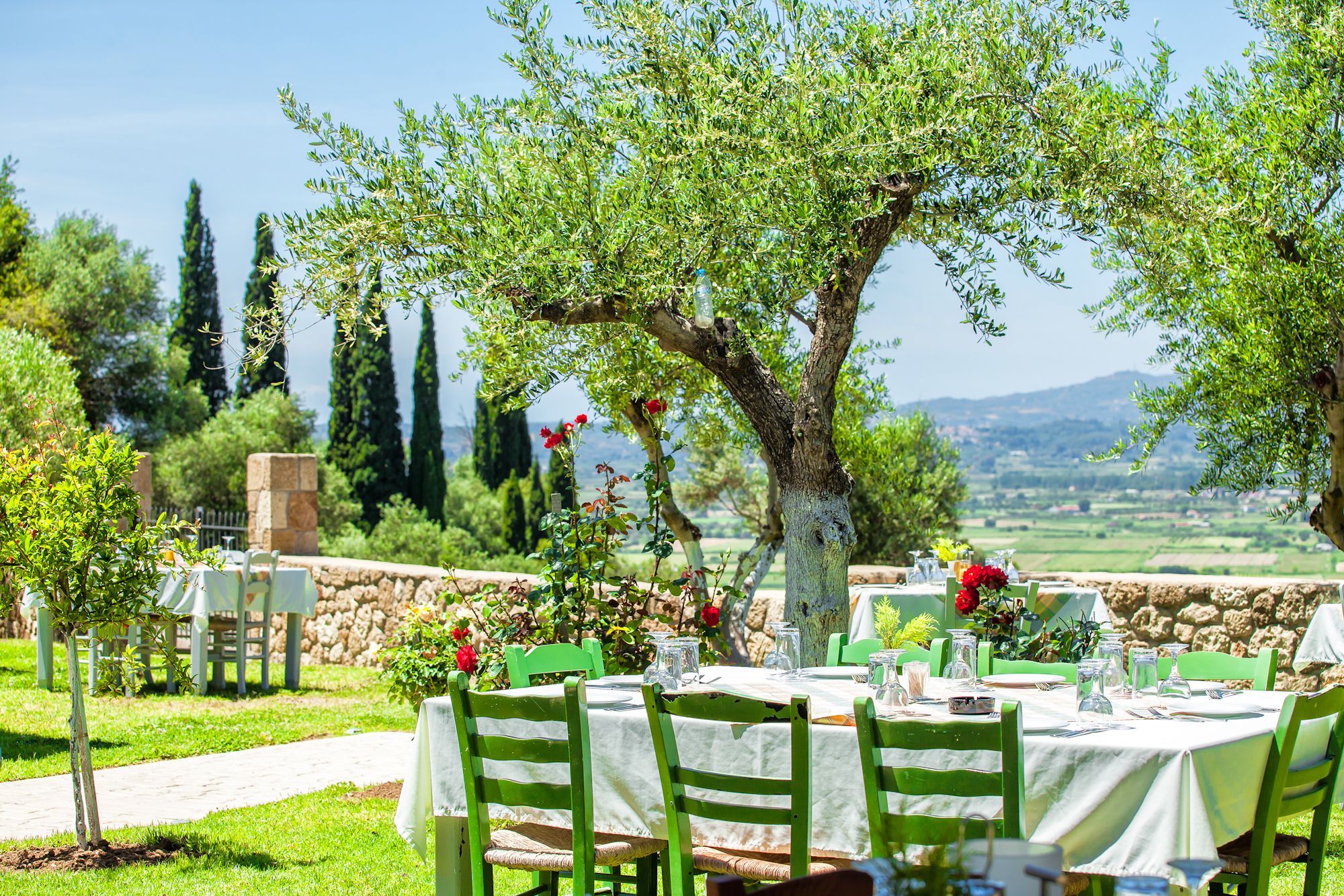 An outdoor dining area with tables set for a meal, surrounded by greenery and a stone wall.