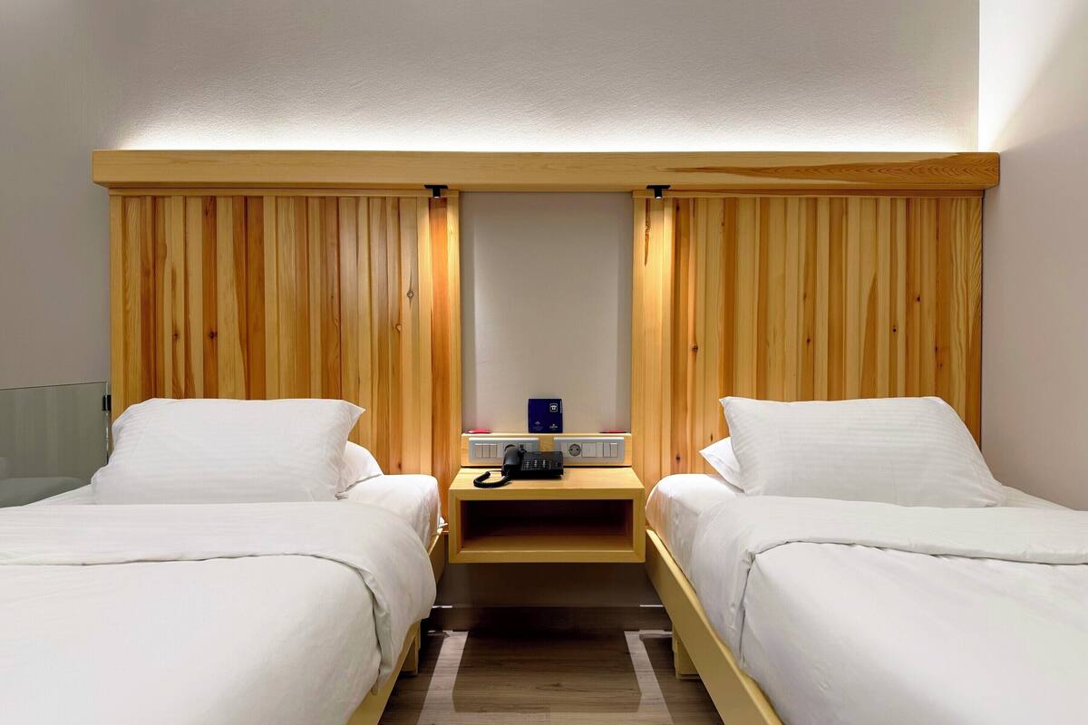 Habitación de hotel con dos camas, paneles de cabecero de madera, una mesita de noche con teléfono y radio, y una lámpara sobre las camas.