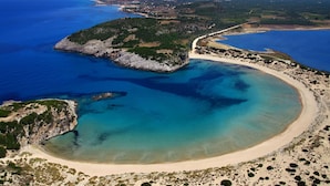 Vue aérienne d’une zone côtière avec une plage de sable, des eaux d’un bleu limpide et une petite île parsemée de formations rocheuses.