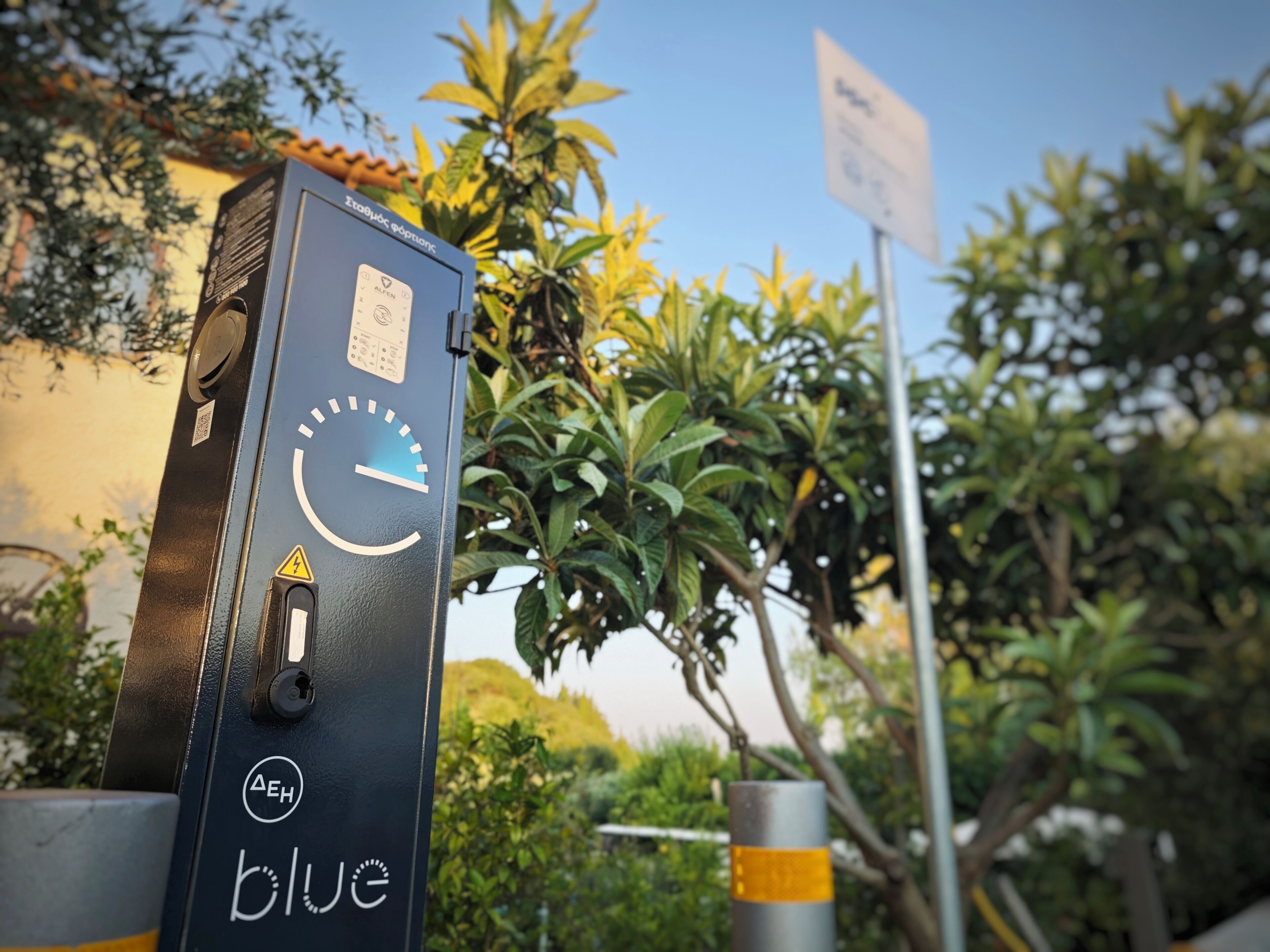 A black electrical box with a dial and a warning sign, surrounded by greenery and a building.