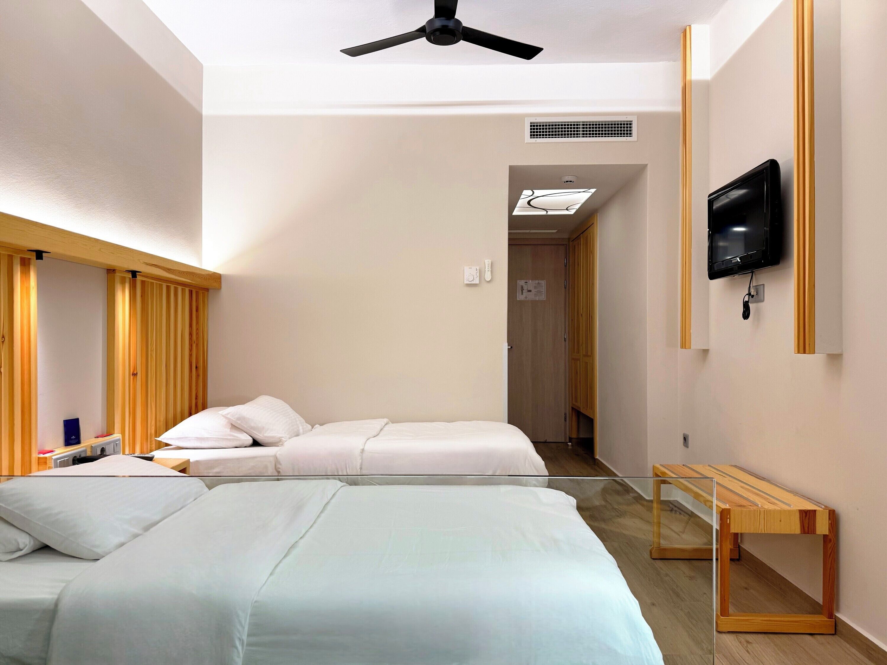 A hotel room with two beds, a ceiling fan, a TV mounted on the wall, and a wooden bench.