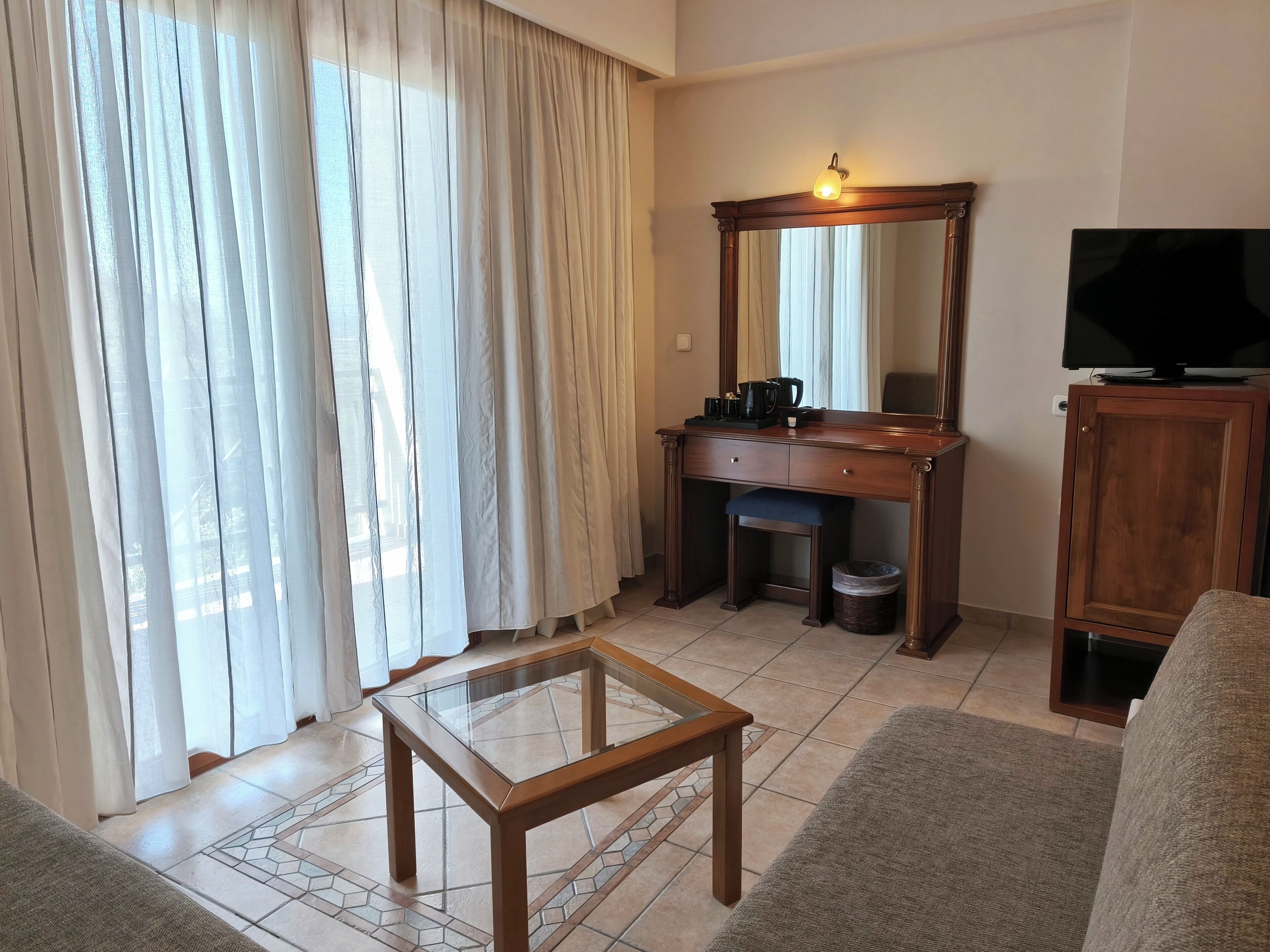 A living room with a glass-top coffee table, a wooden desk with a mirror, a television, and a sofa.