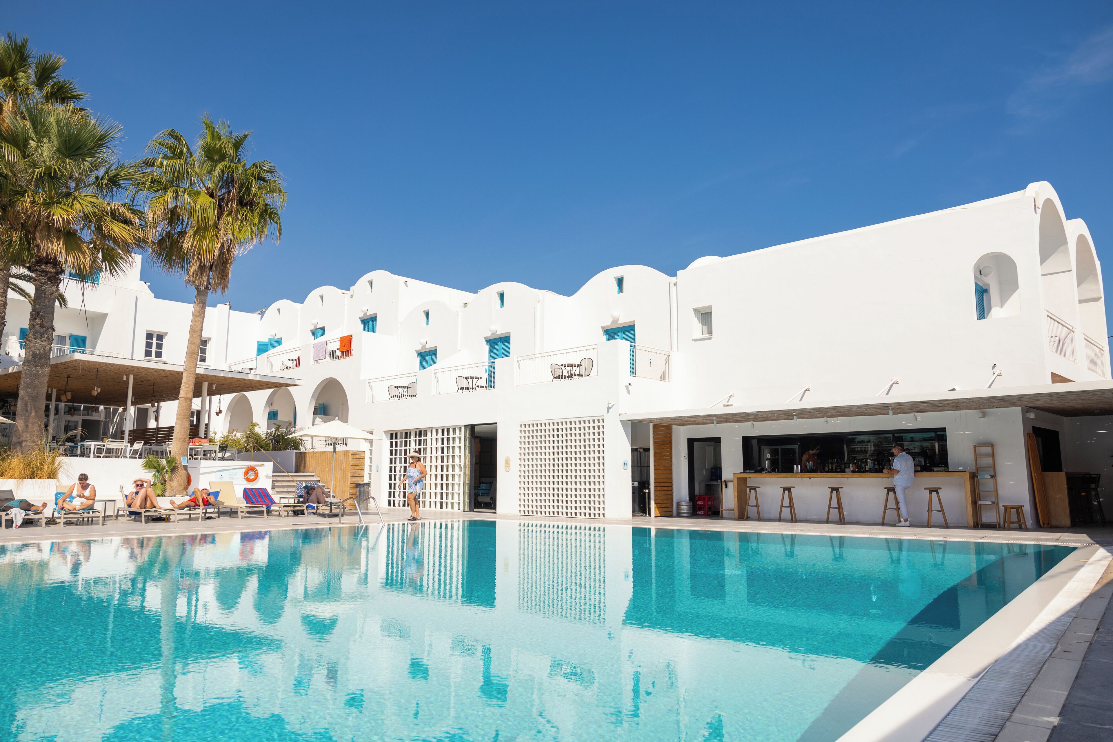 A poolside area with a clear blue pool, white buildings with blue accents, and palm trees.