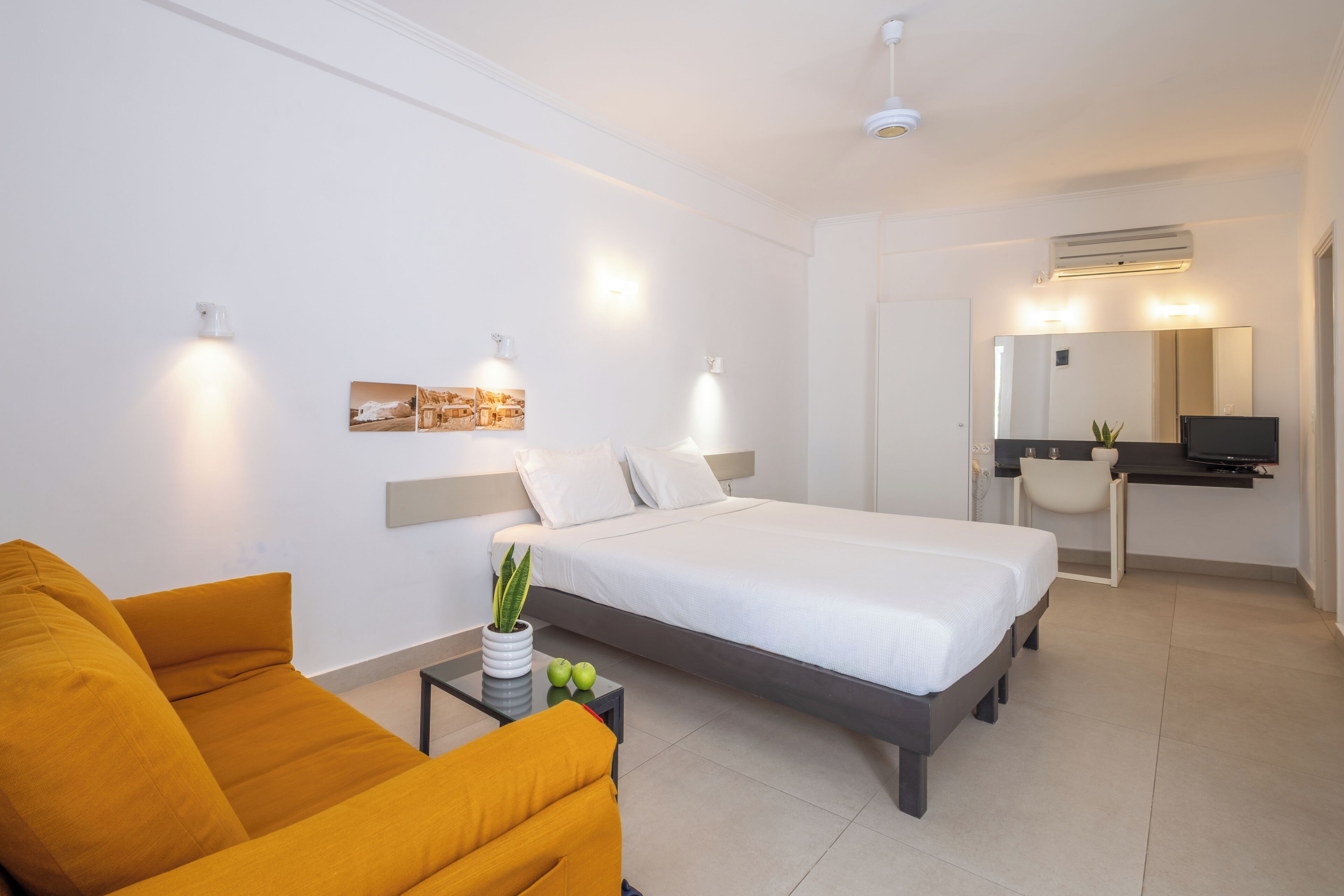 A modern hotel room with a bed, a sofa, a desk with a chair, a TV, and a mirror.