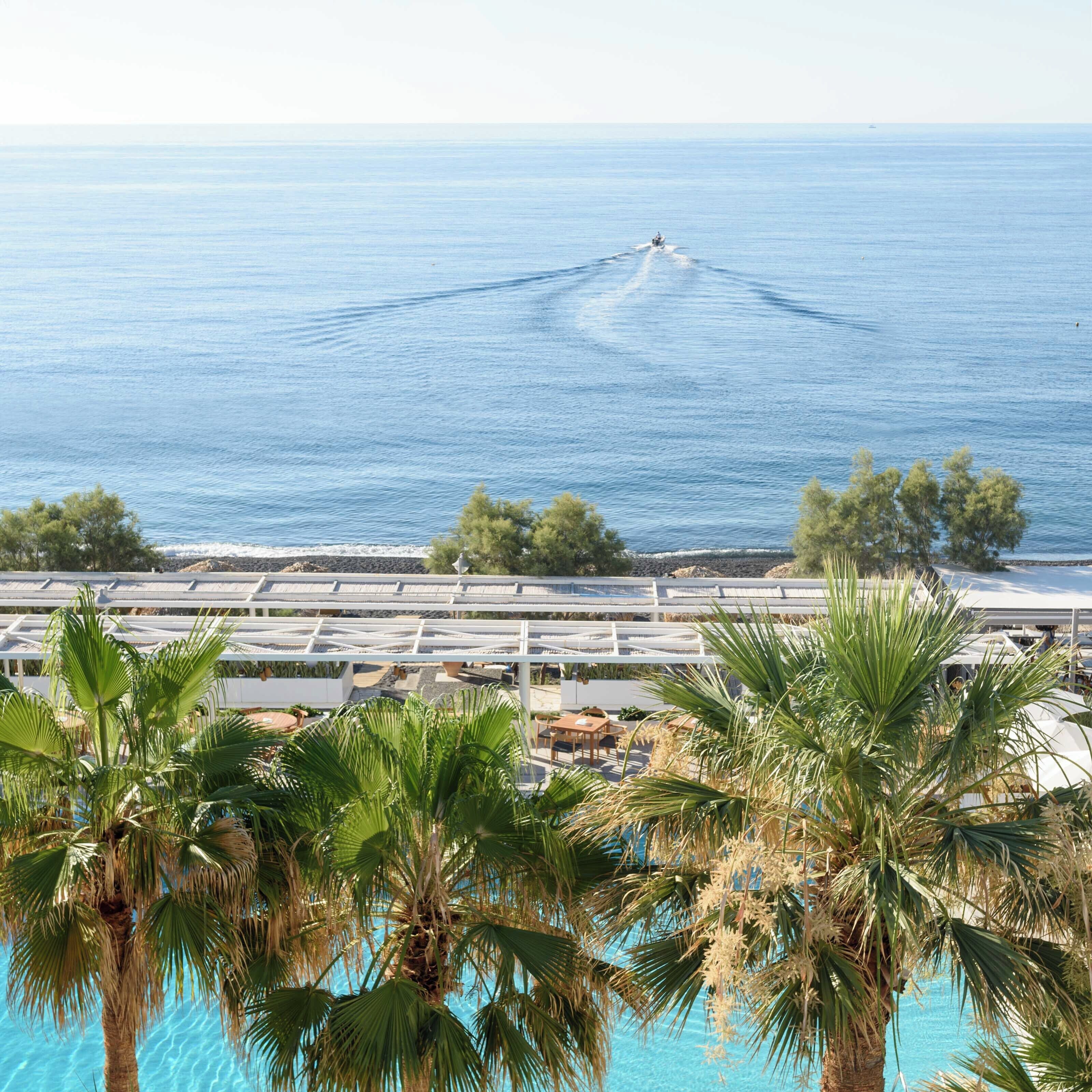 A coastal view with a clear sky, a boat creating a wake in the water, and a beachfront area with palm trees and a pool.