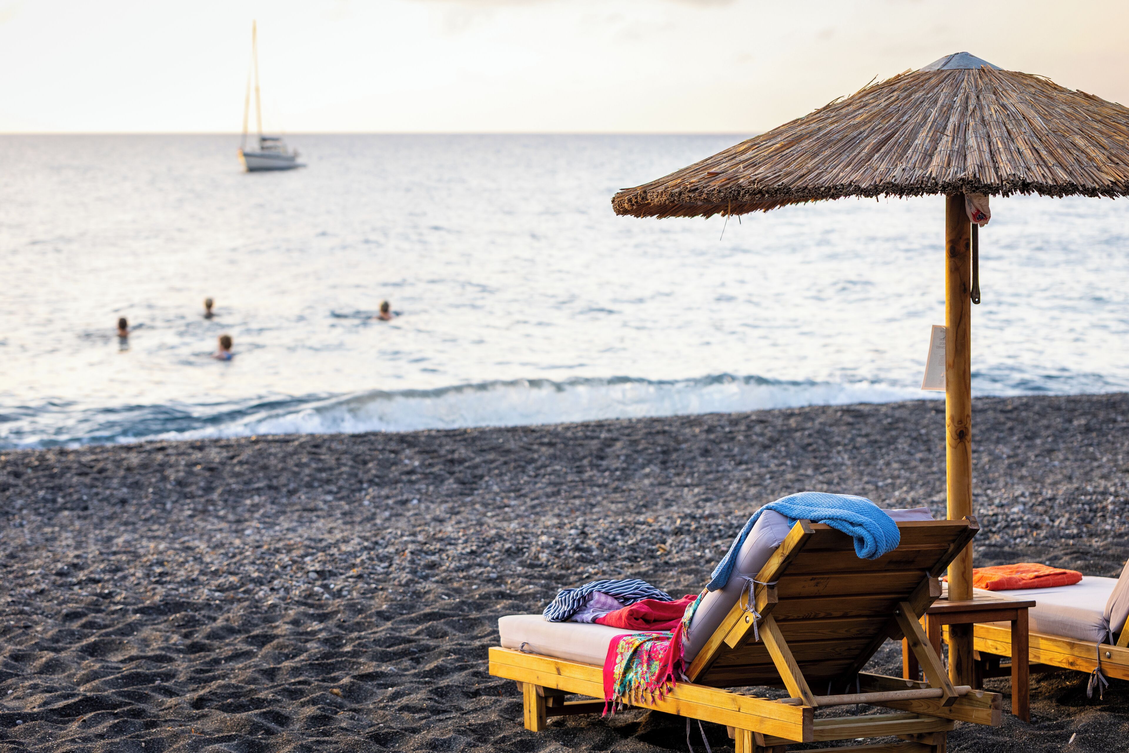 A beach with two wooden lounge chairs under a thatched umbrella, a wooden table, and a view of the sea with a sailboat in the distance.