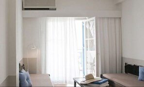 A hotel room with a bed, a table with a hat and folded clothes, a lamp, and a window with curtains. - Kamari Beach Hotel (Santorini)