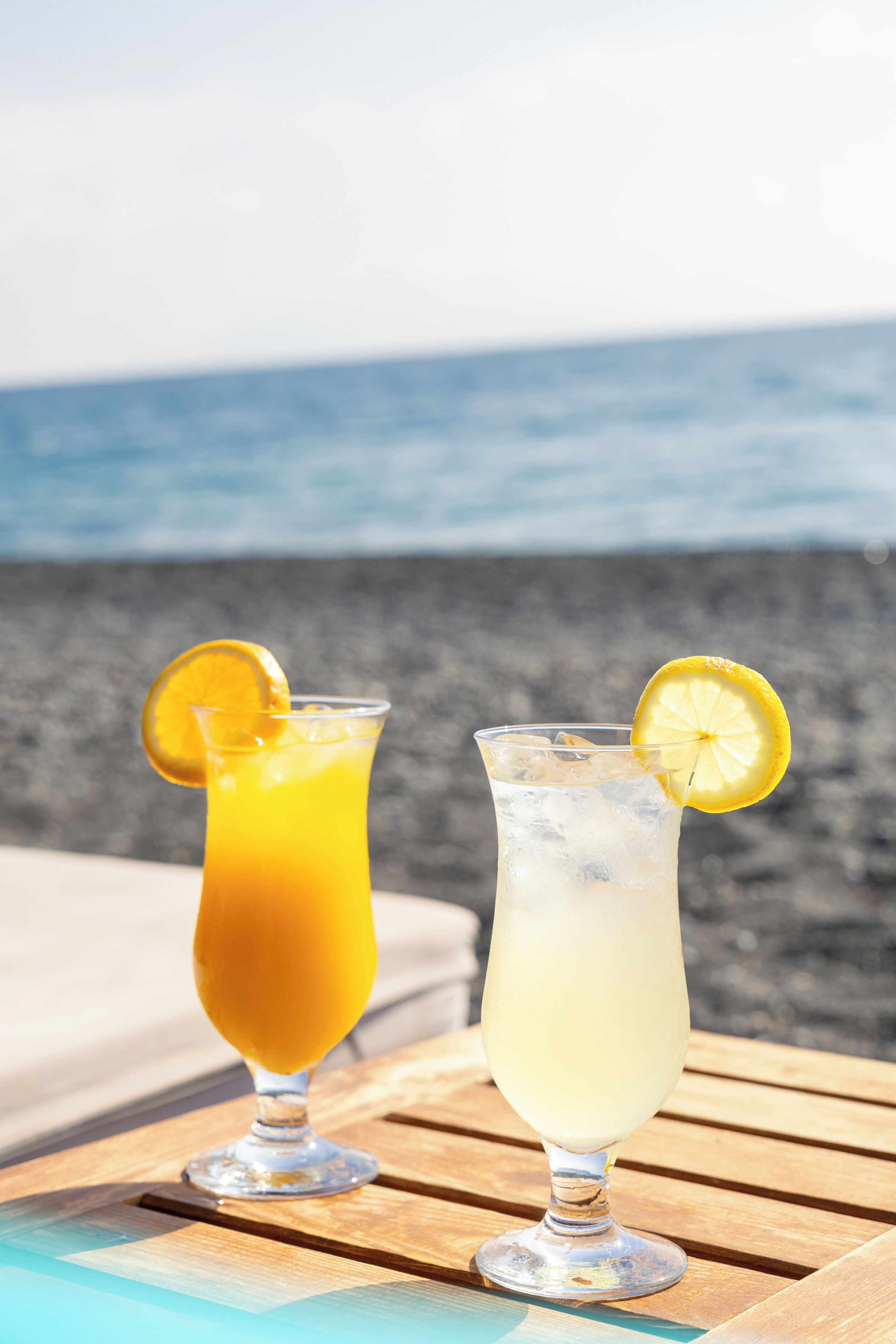 Two drinks on a wooden table with a beach and ocean in the background.