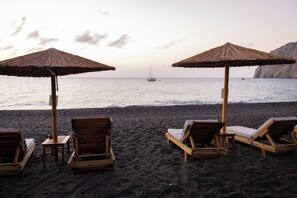 A beach with wooden lounge chairs and thatched umbrellas, a sailboat in the distance, and a rocky cliff in the background. - Kamari Beach Hotel (Santorini)
