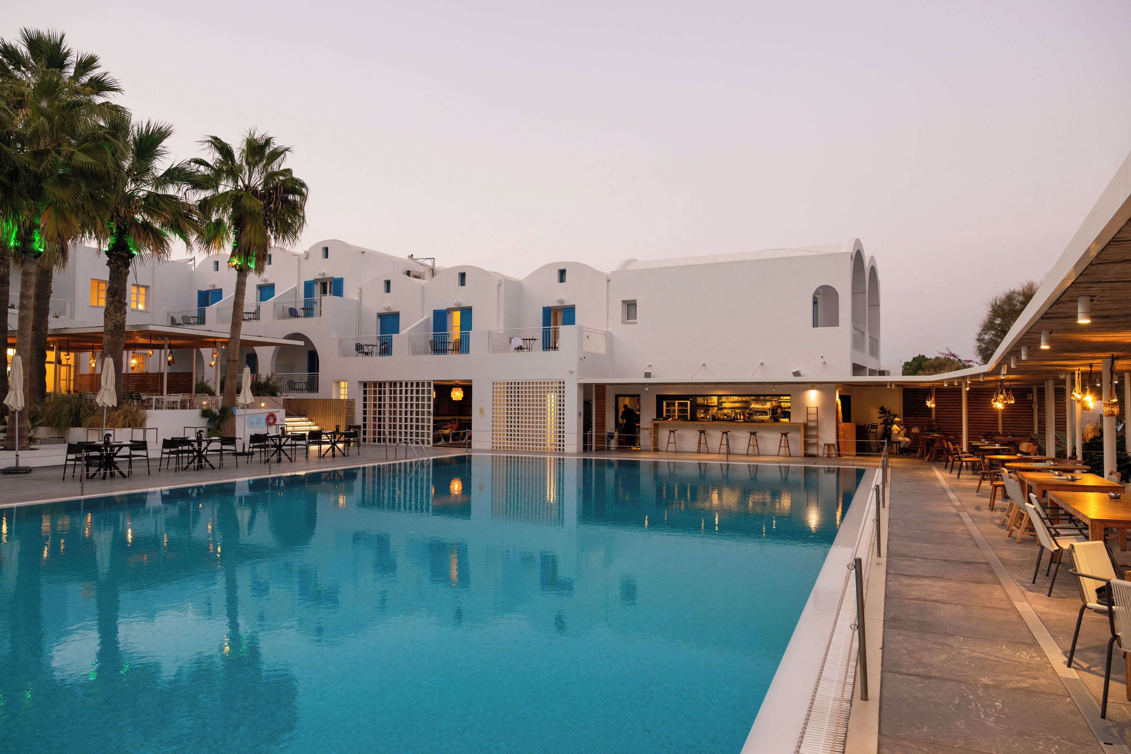 A swimming pool with surrounding tables and chairs, a bar area, and a building with multiple balconies.