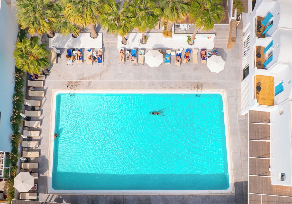 Una piscina en la azotea con sillones y sombrillas, rodeada de palmeras y un edificio con balcones.