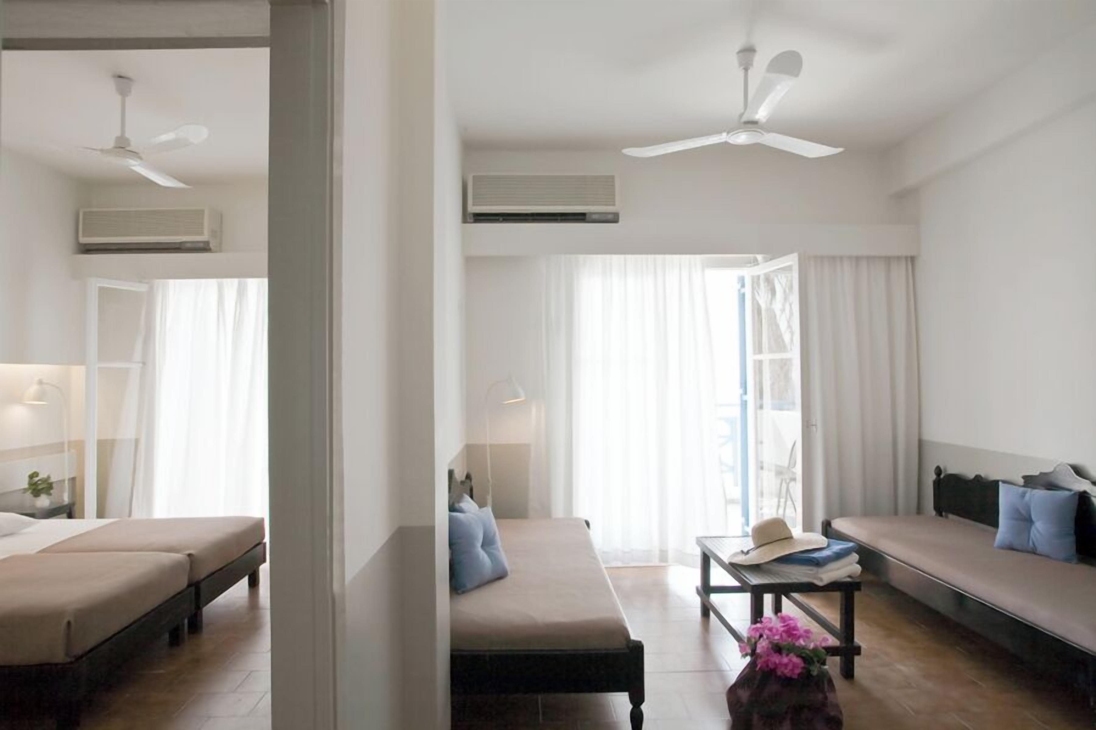 Ein Hotelzimmer mit zwei Betten, einem Schlafsofa, einem Tisch mit Hut und Büchern sowie einem Deckenventilator.