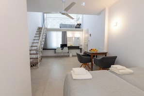 A modern hotel room with a bed, a desk, a chair, a small table with fruit, a mirror, and a staircase. - Kamari Beach Hotel (Santorini)