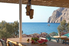 A wooden table with fruit and a hanging basket, overlooking a coastal view with boats and cliffs. - Kamari Beach Hotel (Santorini)
