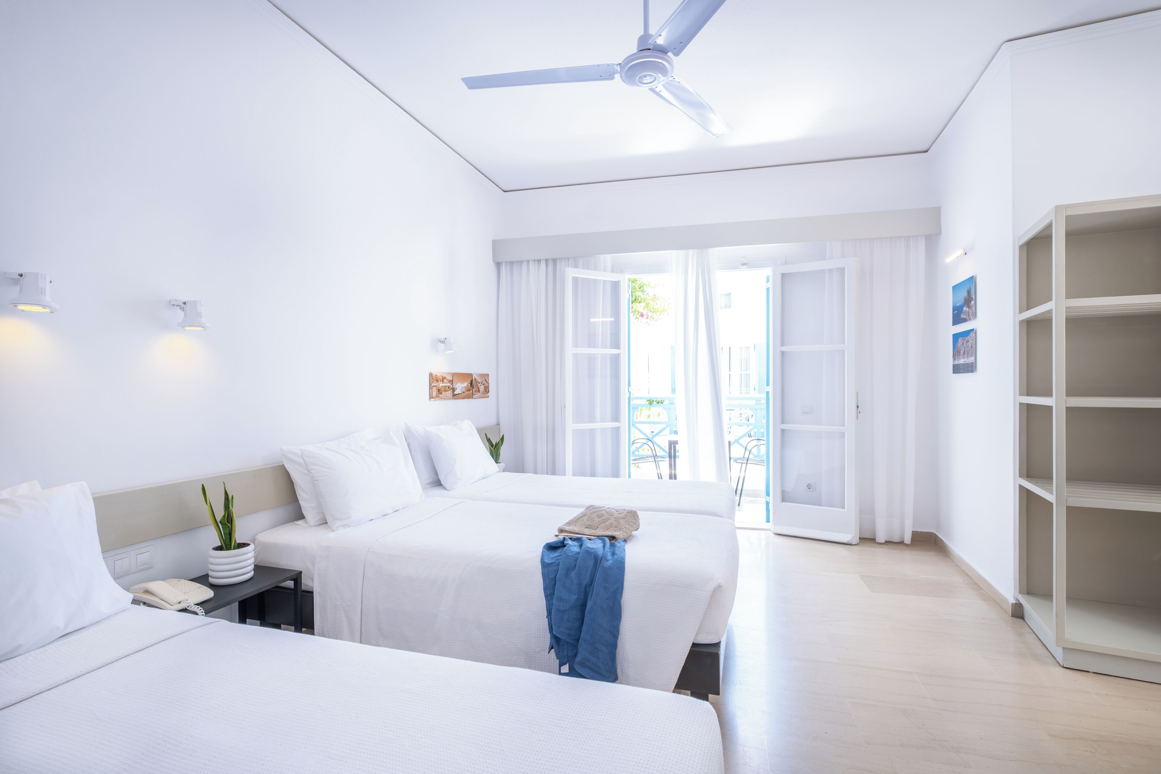 Une chambre d’hôtel avec deux lits, un ventilateur de plafond, une petite table avec une plante, une lampe de chevet et une vue sur l’extérieur.