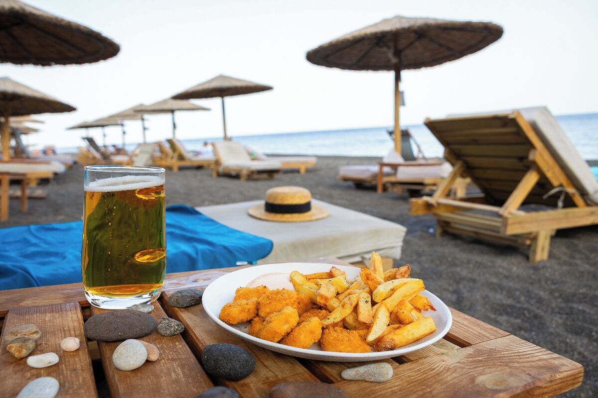 Escena de playa con una mesa que presenta un plato de comida y una jarra de cerveza, rodeada de sillas y sombrillas de playa.