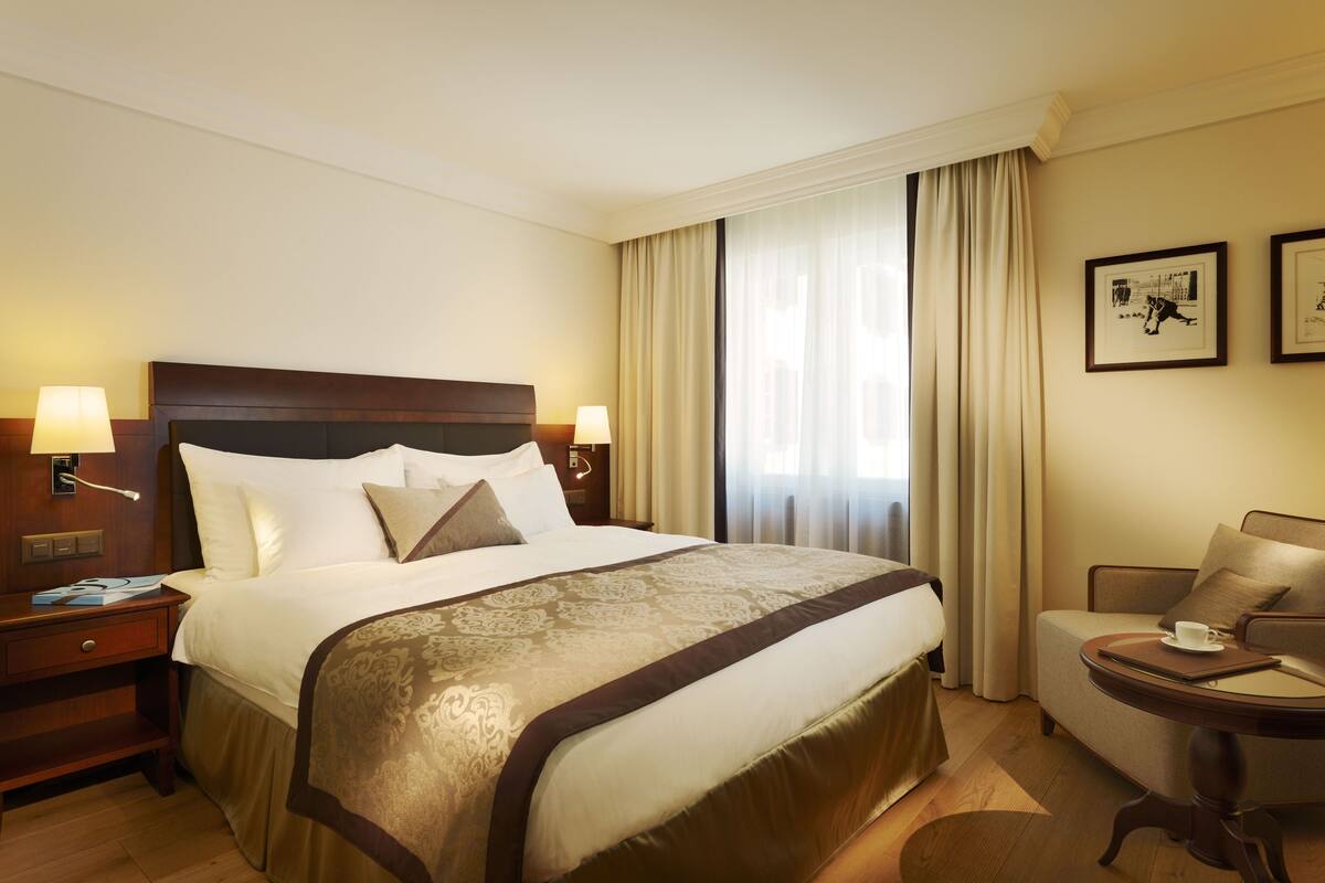 A hotel room with a large bed, two bedside tables with lamps, a small round table with a cup and saucer, and a chair.
